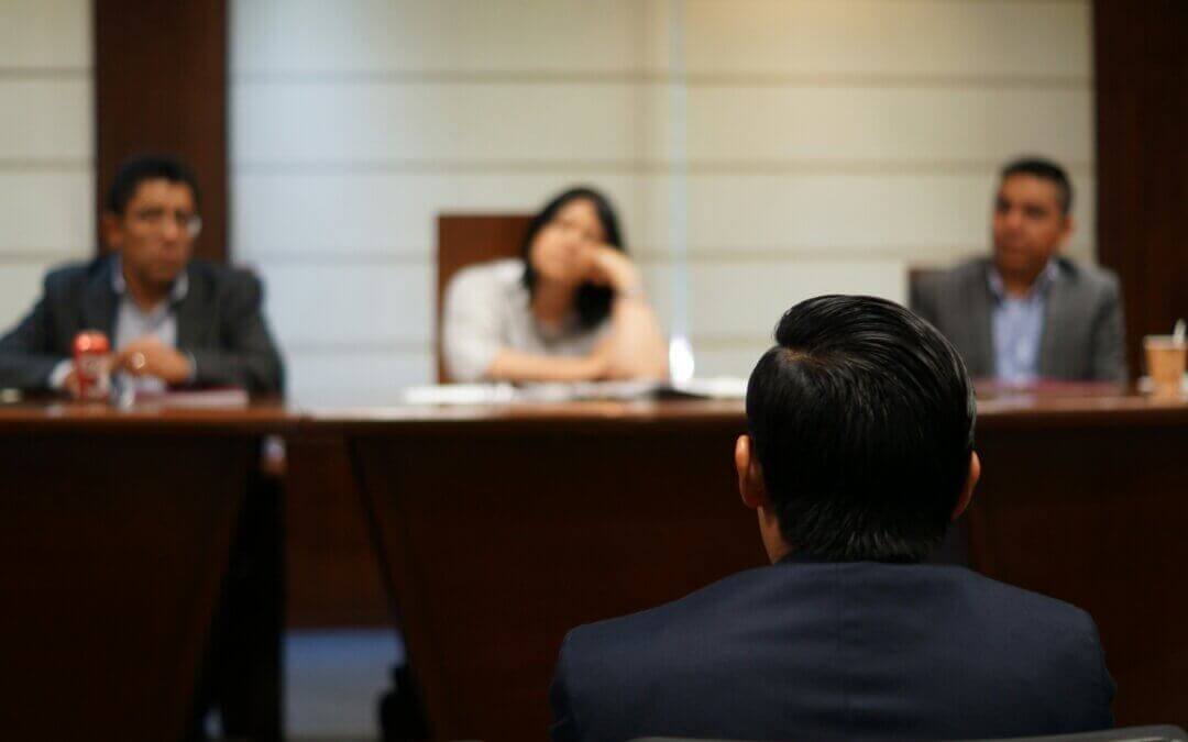 A person sits facing three individuals across a conference table in a formal meeting room. The three people are looking toward the person, with one appearing to listen intently and another resting their chin on their hand, as if pondering whether you can be charged without being arrested.