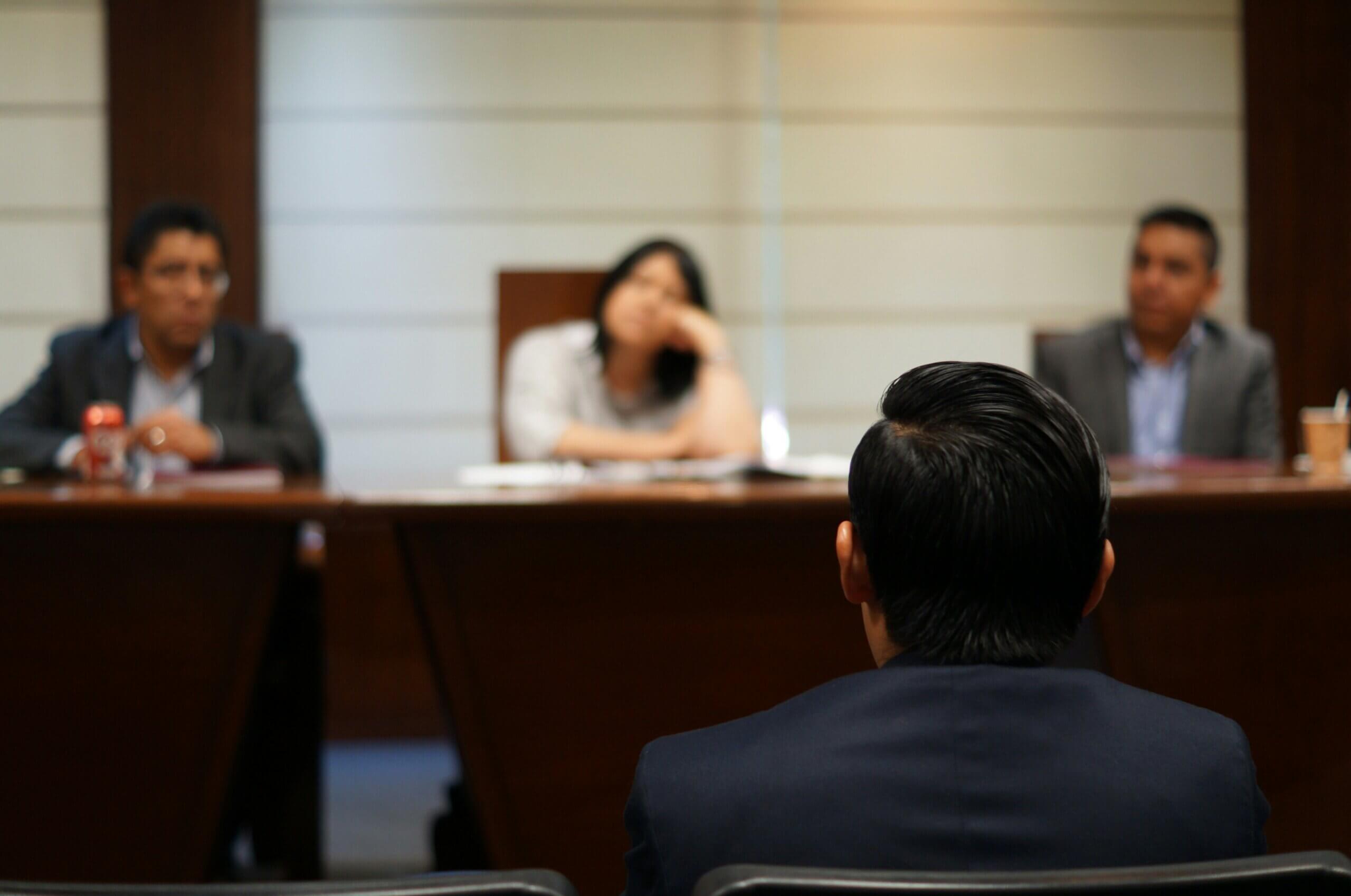 A person sits facing three individuals across a conference table in a formal meeting room. The three people are looking toward the person, with one appearing to listen intently and another resting their chin on their hand, as if pondering whether you can be charged without being arrested.