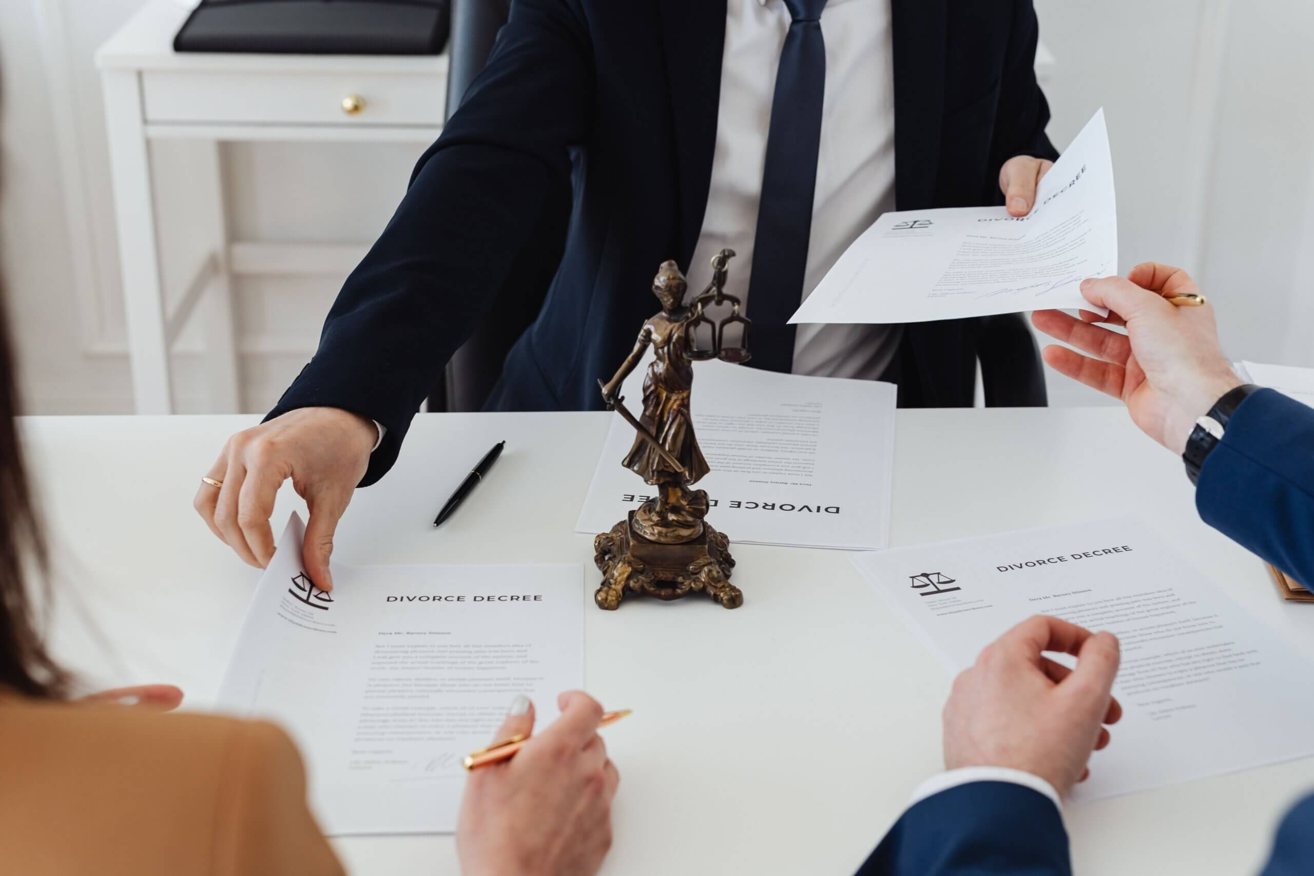 Three people in suits exchange divorce decree documents at a table with a Lady Justice statue in the center, subtly reflecting the balance of justice seen in Arkansas Self-Defense Law. One person holds a pen, ready to sign. Papers and a pen are on the white table.