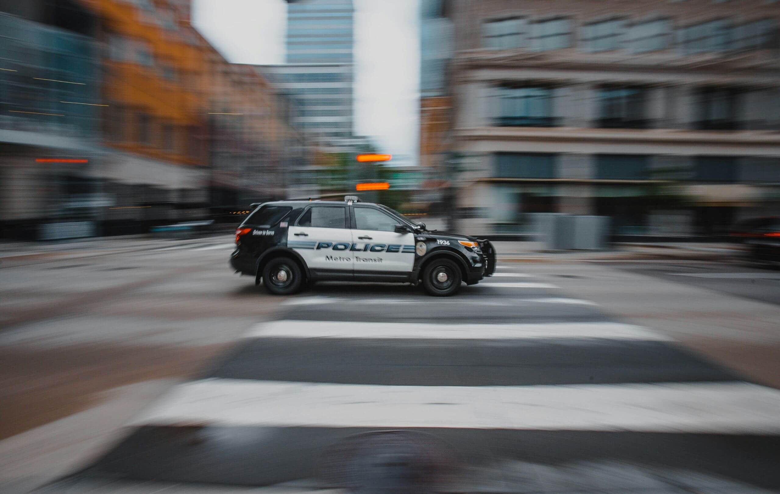 What Makes a Search or Seizure in Arkansas Illegal A police SUV speeds through a city intersection, its markings reading Police and Metro Transit, possibly responding to a search or seizure. Surrounding buildings blur with motion, while a crosswalk lines the foreground.