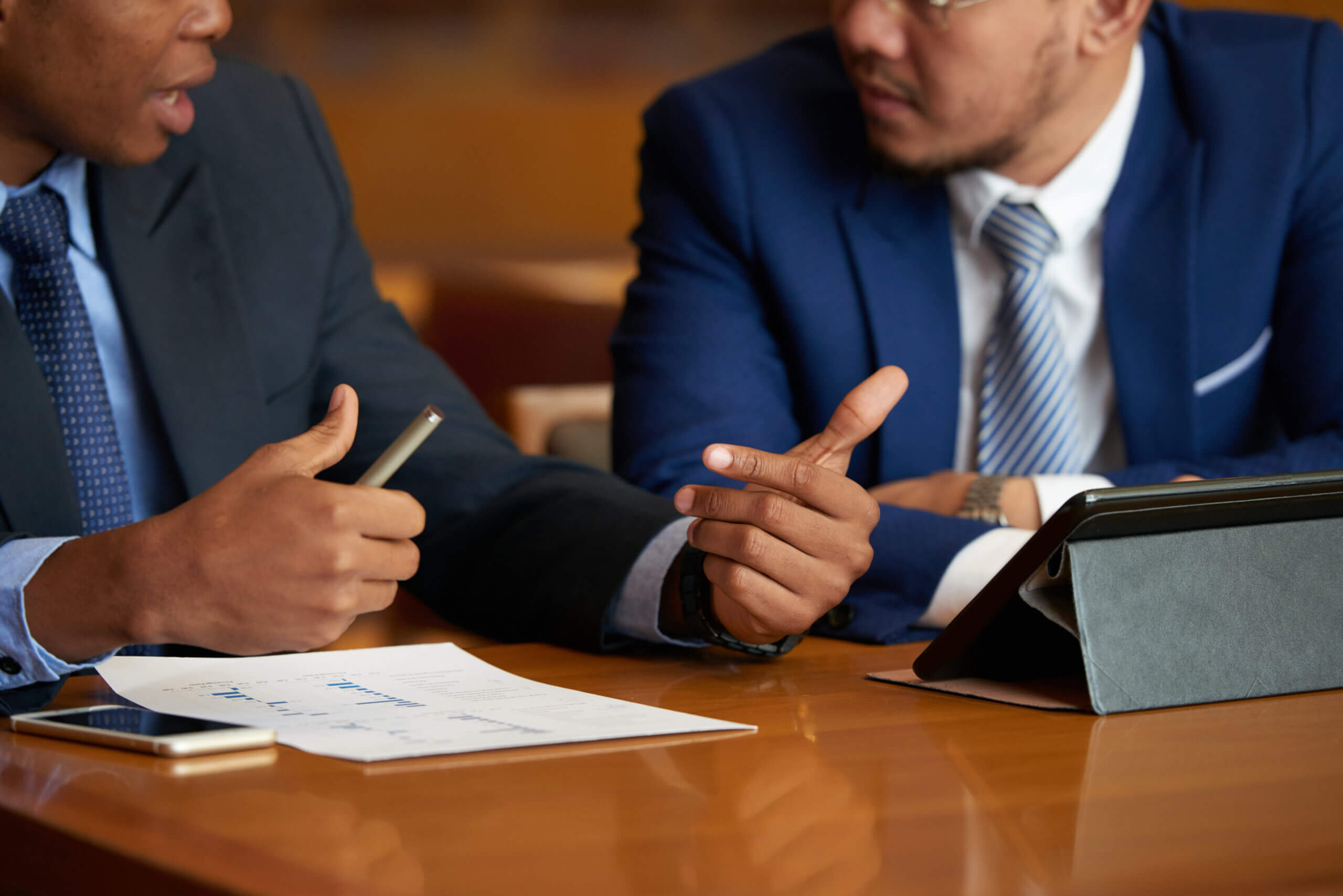 Two men in suits sit at a table, discussing documents and charts with a tablet and smartphone nearby. One gestures with a pen while the other listens attentively, as if reviewing data from a recent witness testimony.