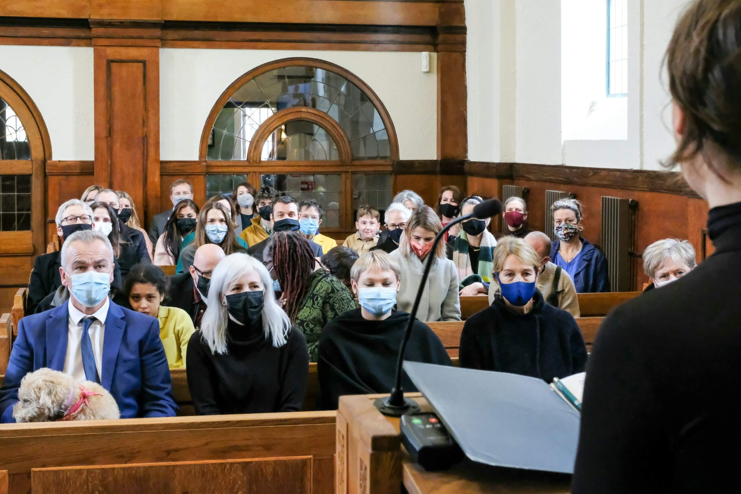A group of people wearing face masks sit on wooden pews in a church, listening attentively to a speaker delivering witness testimony. The room features large windows and wood paneling, creating a warm and solemn atmosphere.