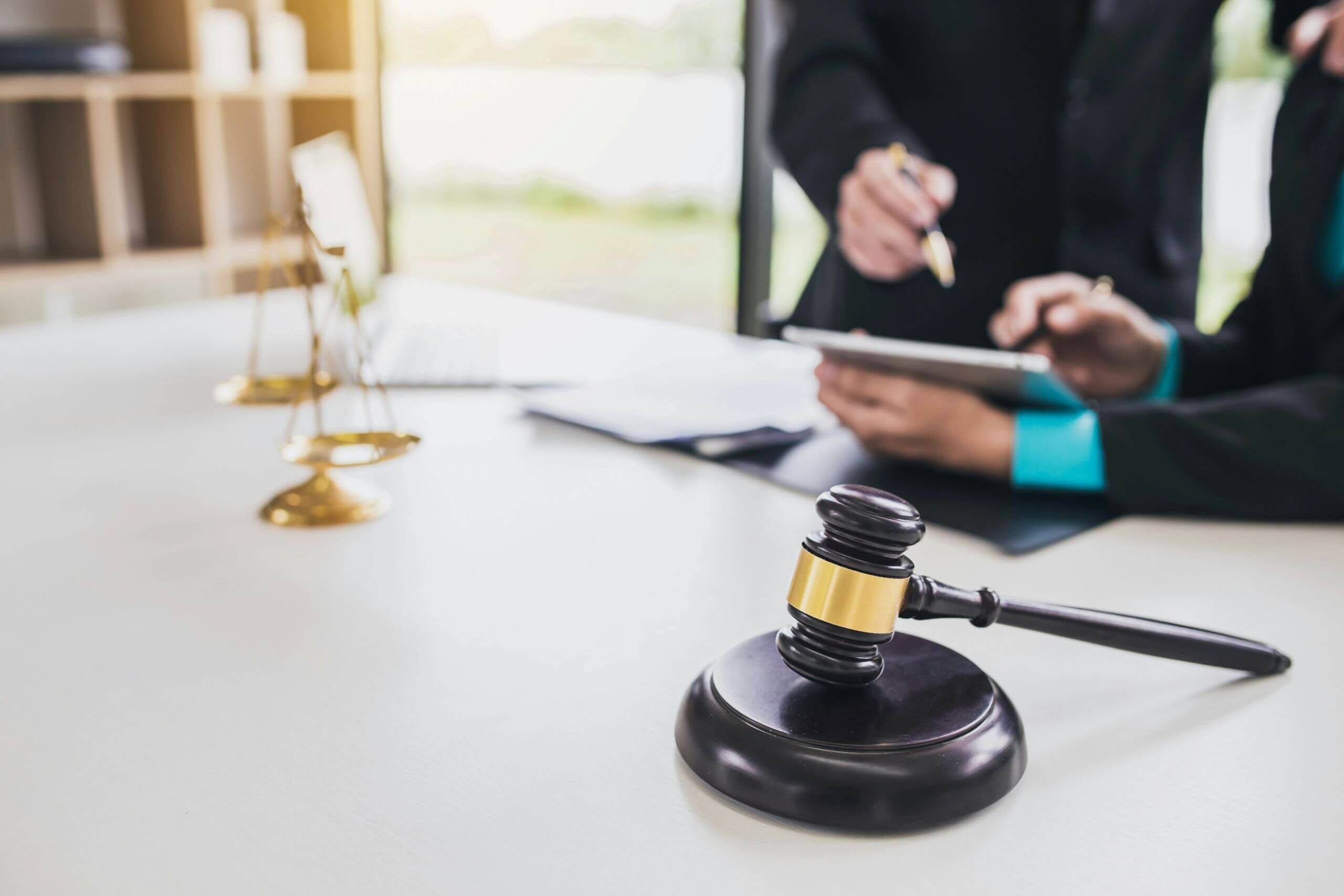 jury trials A wooden gavel and scales of justice sit on a desk, while two people in business attire discuss documents and use a tablet in the background, suggesting a law office setting focused on jury trials.