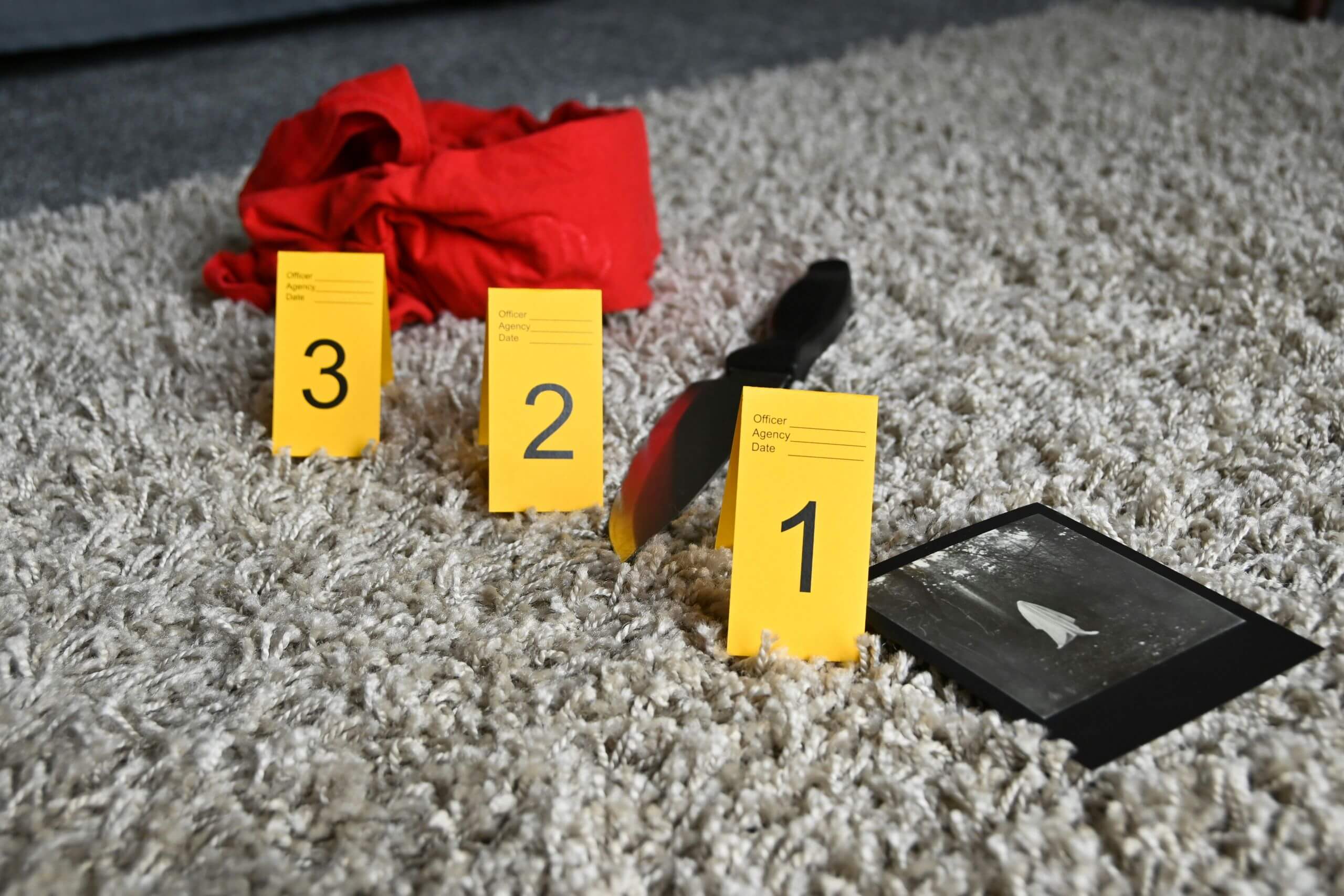 A crime scene with three yellow evidence markers numbered 1 to 3 highlights crucial physical evidence: a knife, a red cloth, and a black-and-white photo scattered across a light-colored carpet.