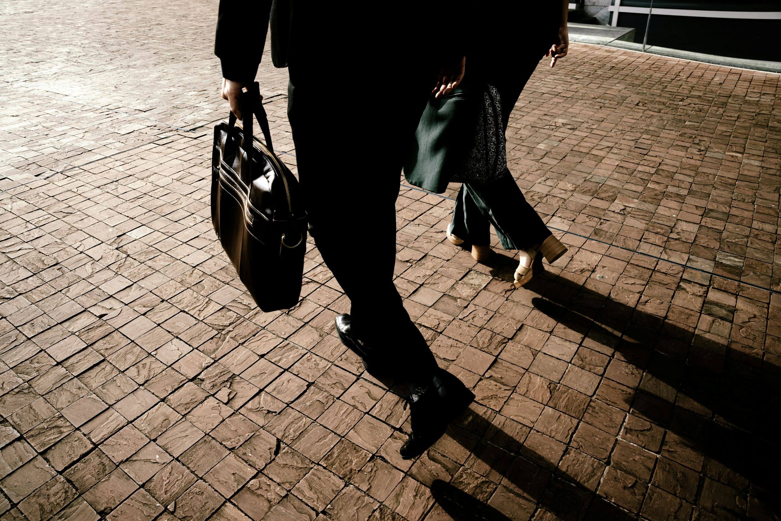 Two people in business attire walk on a sunlit brick pavement. One carries a briefcase, and their long shadows extend across the ground. Only their lower bodies are visible.