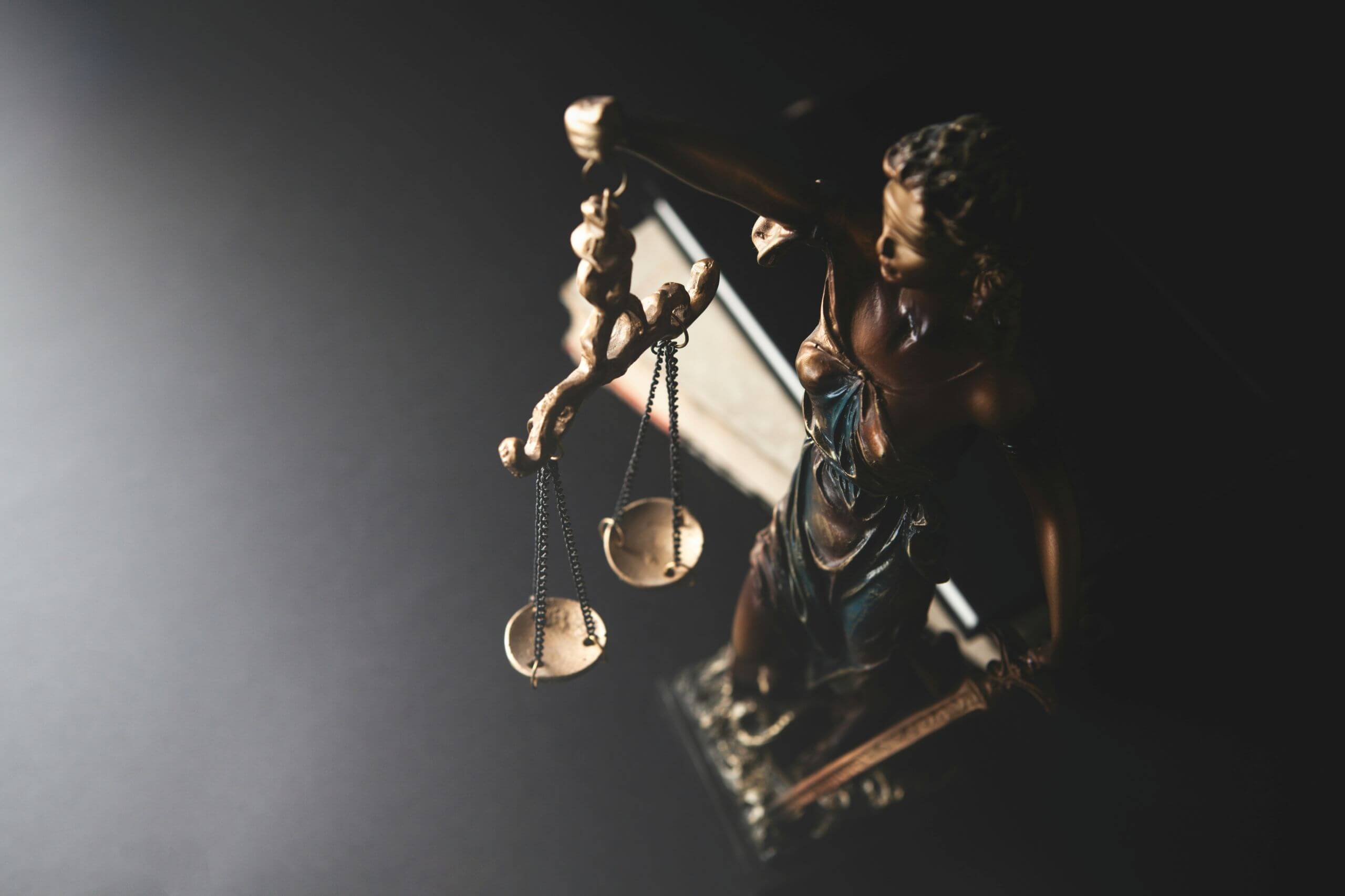 A bronze statue of Lady Justice holding scales and a sword is seen from above, with dramatic lighting casting shadows on a dark background.