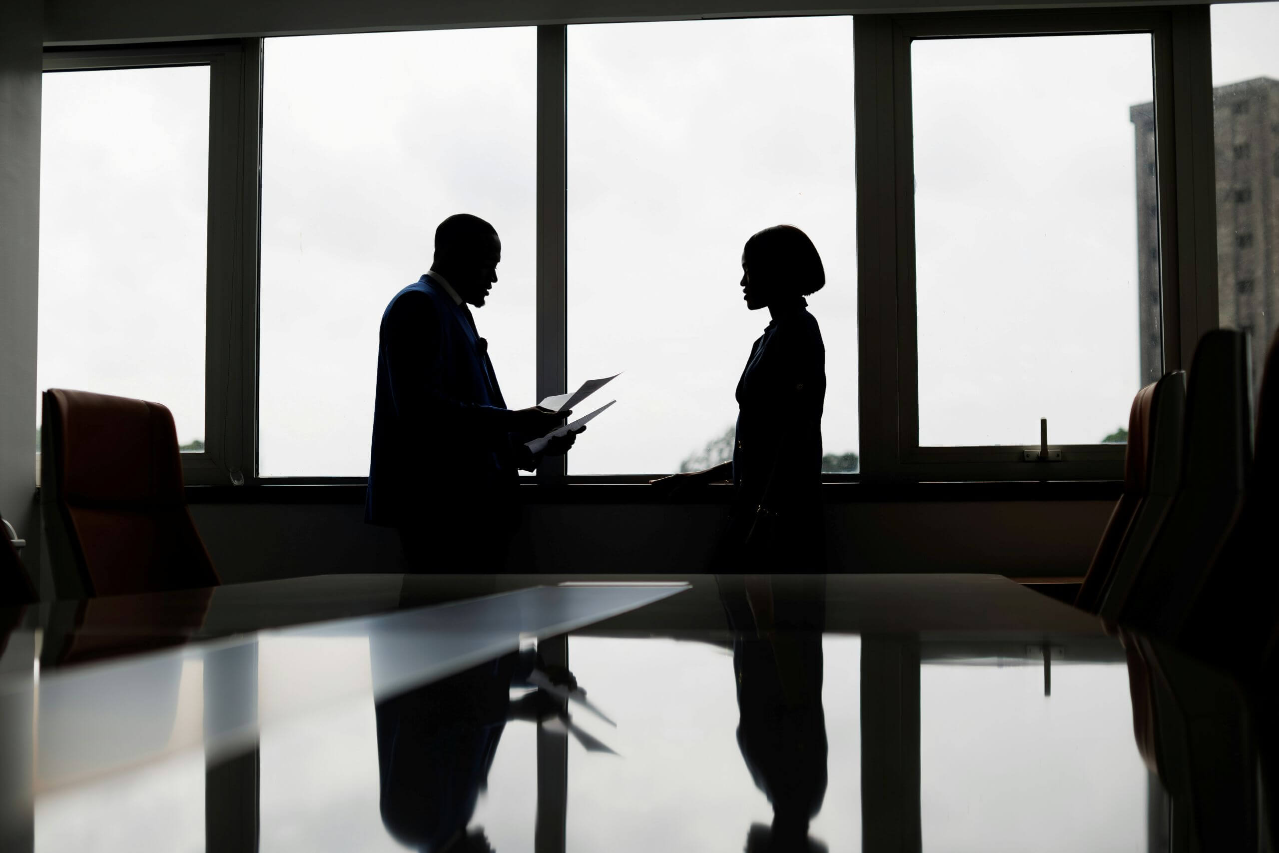 What Is Entrapment and How Is It Proven in Arkansas Two people in business attire are silhouetted, standing and talking in front of large office windows, with papers in hand. The scene is reflected on a glossy table surface.