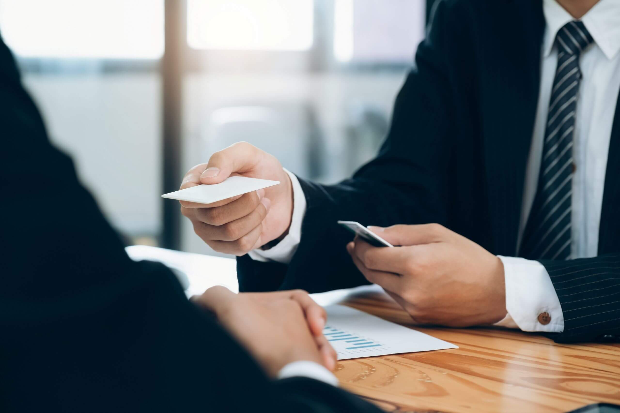 criminal negligence Two people in business suits sit at a table. One is handing over a business card while the other holds papers with a chart. The setting appears to be an office or meeting room.