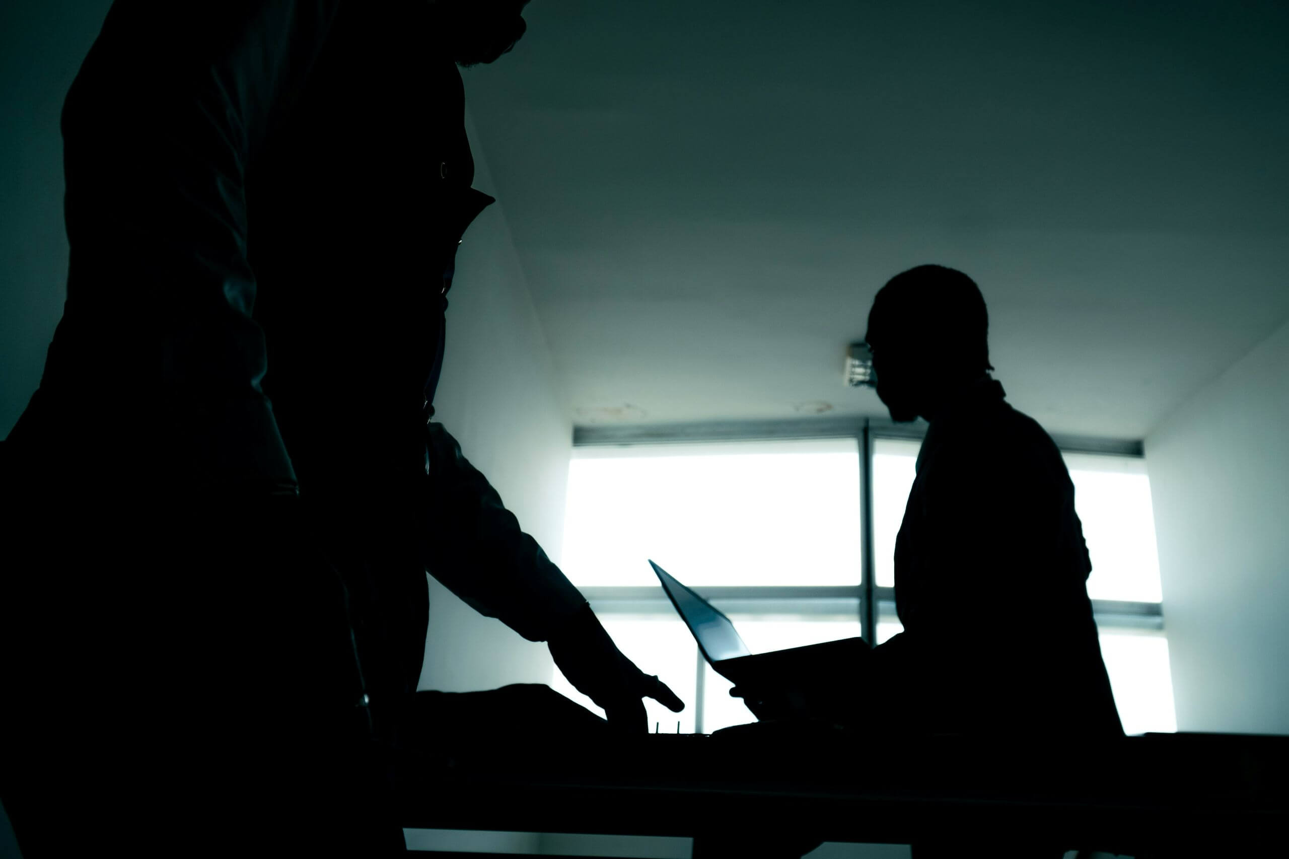 Silhouettes of two people in a dimly lit room, one sitting with a laptop and the other standing, with light coming from a window in the background.